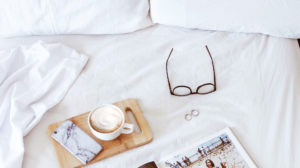 A hotel bed with white linens including a magazine and a cup of coffee on the bed